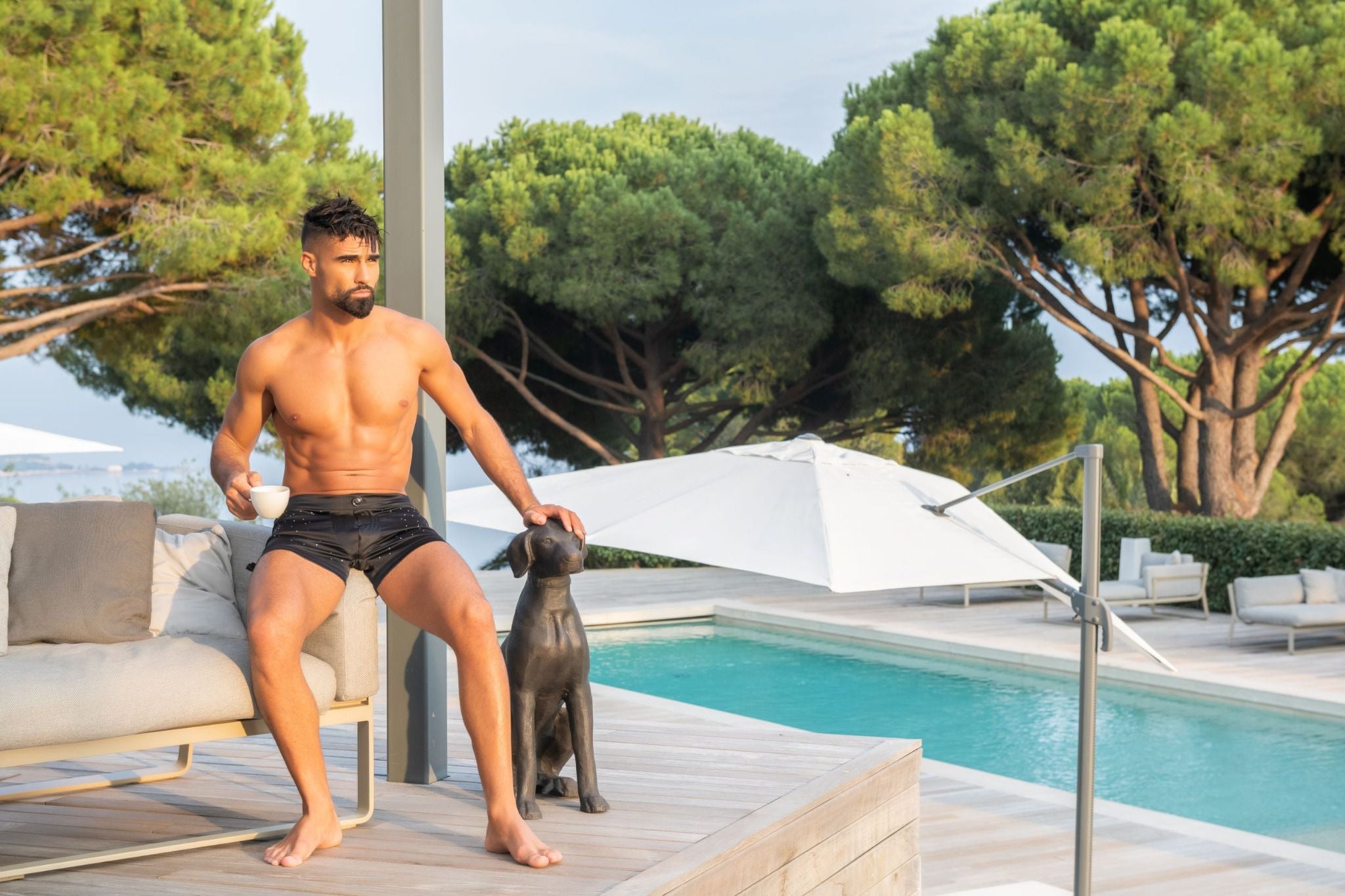 Modèle relaxant au bord d'une piscine luxueuse avec un chien, portant le Boxer Bijou DIAMOND en soie noire, mettant en avant la ceinture élastique soyeuse et les boutons brillants.