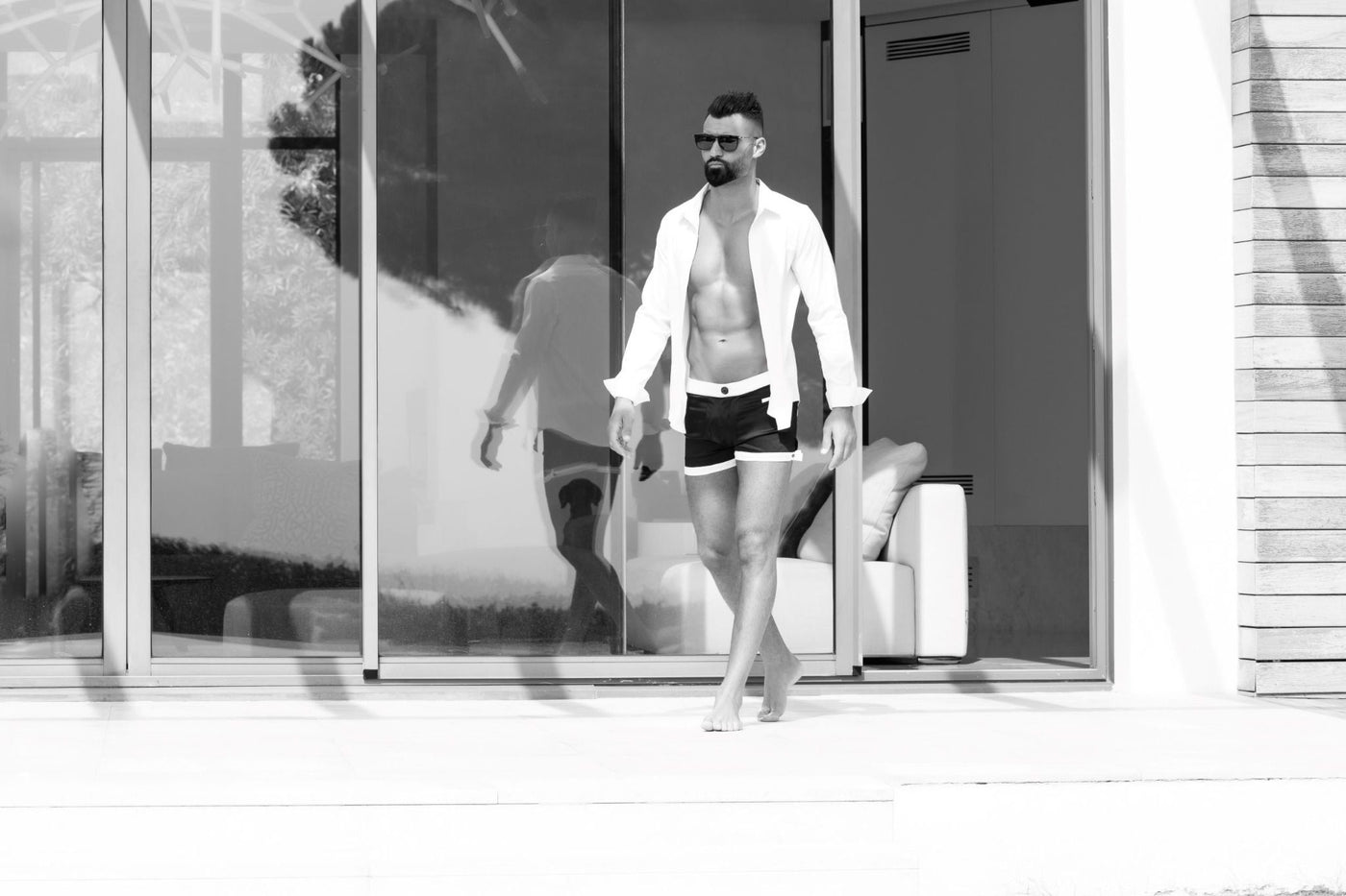 Image en noir et blanc d'un homme marchant en boxer The Classique, devant une villa contemporaine, capturant l'essence de la classe avec un design épuré et des matériaux de qualité.