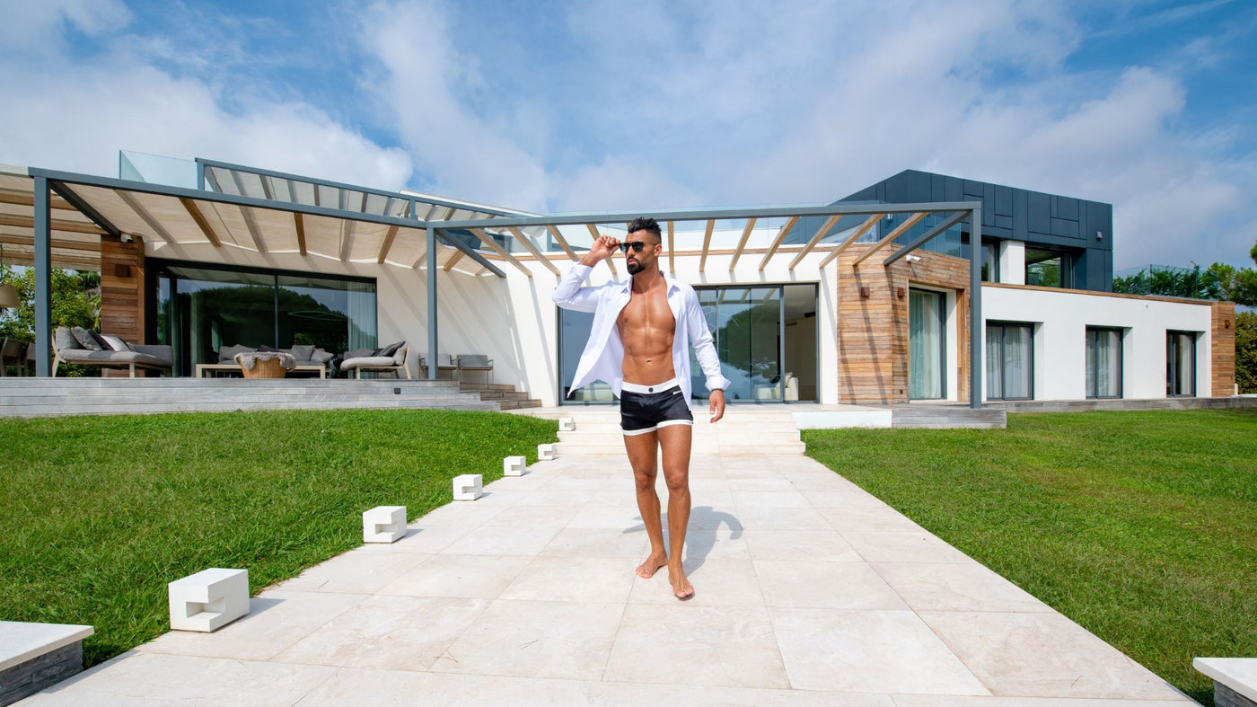 Modèle masculin décontracté ajustant ses lunettes de soleil en marchant près d'une villa moderne, vêtu du boxer The Classique qui contraste avec son style décontracté chic.