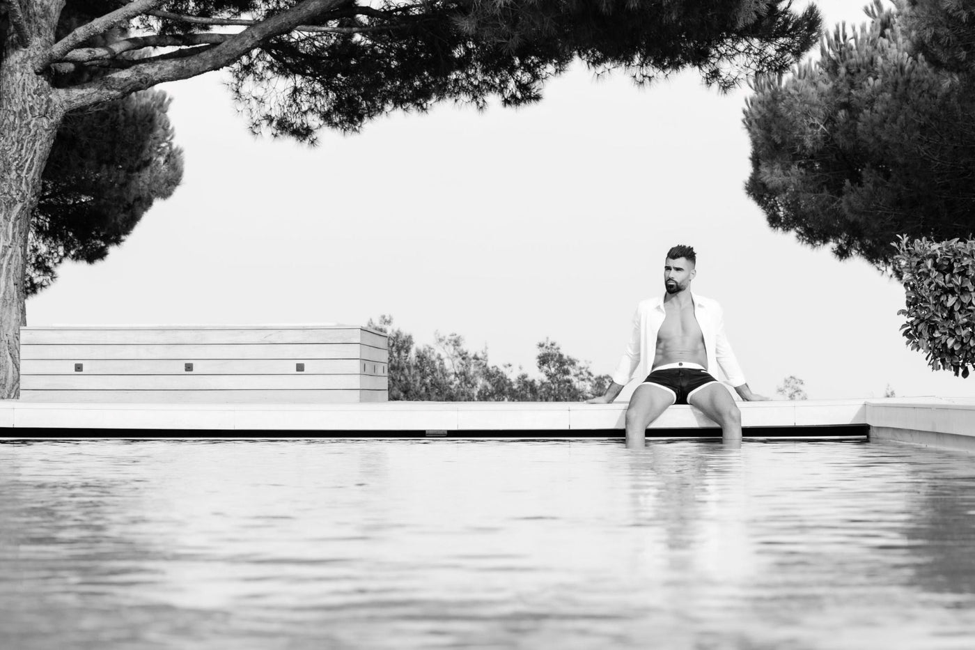 Image en noir et blanc d'un homme assis au bord d'une piscine, reflétant l'élégance intemporelle du boxer The Classique, accentuée par la ceinture élastique en soie blanche.