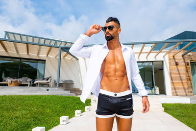 Modèle masculin en The Classique boxer, touchant légèrement ses lunettes de soleil, évoquant une esthétique luxueuse avec la villa moderne en arrière-plan.